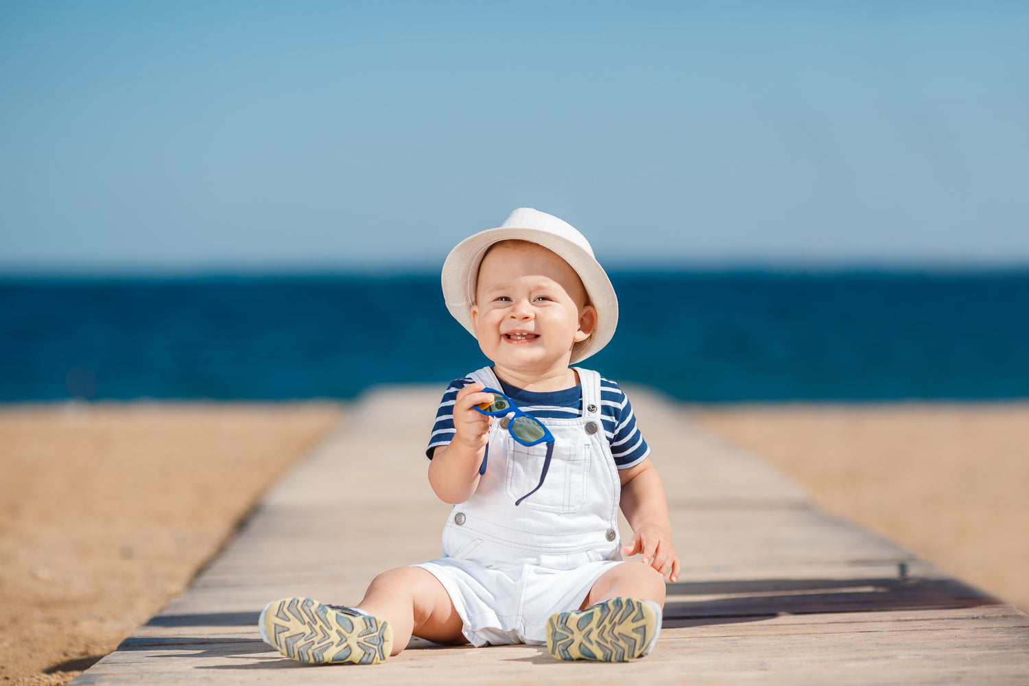 Bébé assis sur un ponton en bois à la plage, portant un chapeau blanc et tenant des lunettes de soleil, avec la mer en arrière-plan